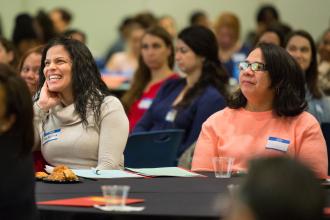 Two women listen in on a presentation while one laughs at something said. 