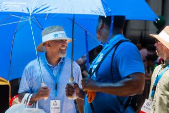Three men talking while walking through the parade. The man on the left and the man on the right are smiling and wearing tan fedora hats The man in the middle is turned to the man to the left. 