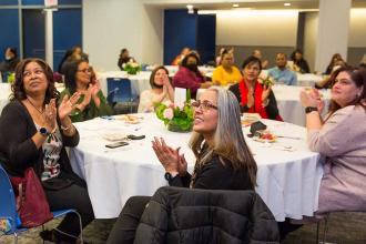 A group of women at a table clap for a speaker.