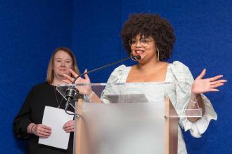 A woman speaks at the podium with her hands stretched outward