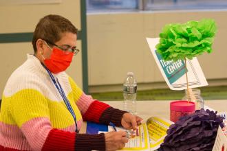 A woman wearing a red mask sits at a table and takes notes 