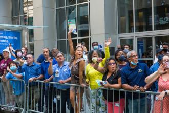 Several people on the sidewalk behind a barricade are all cheering and smiling