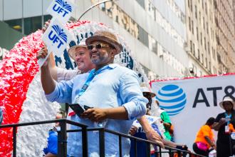 Two men on the float, both wearing fedora hats and one wearing sunglasses, smiling on the float. 