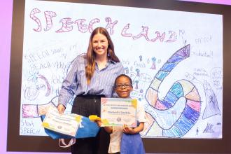 Speech teacher Jessica McGee of PS 78 in the Bronx and student Malachi beam as they stand in front of an image of the Speech Land game board Malachi drew to illustrate the gains he’s made along his speech therapy journey. 