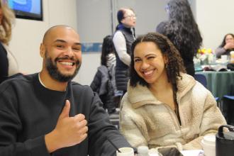 A social worker on the left flashes a "thumbs up" as the social worker next to him smiles. The two social workers are seated at a table during the 2026 UFT Clinicians Appreciation Day event. 