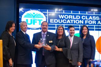 Award winner showing aware surrounded by others in front of an electronic screen