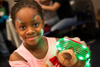 Smiling girl with rainbows painted on her face holding a teddy bear
