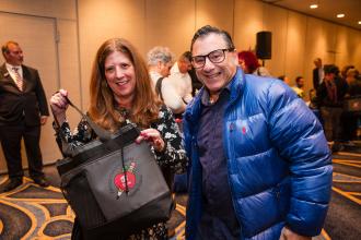 A woman on the left poses for a photo while holding a bag she received at the UFT New Retiree Luncheon while her husband smiles beside her. 
