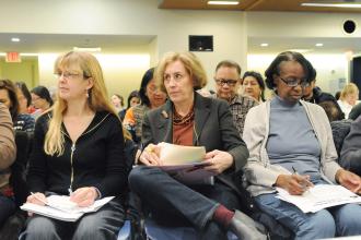 Three women holding papers seated in large group of people