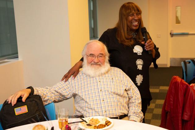 A woman stands next to a man sitting at a table