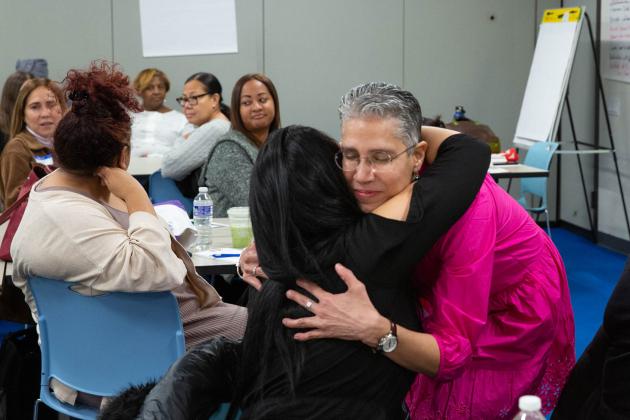 A woman in a pink shirt hugs a woman in a black shirt. 
