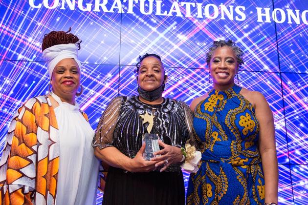 Three women pose for a photo while on stage. The person in the middle is holding a plaque 