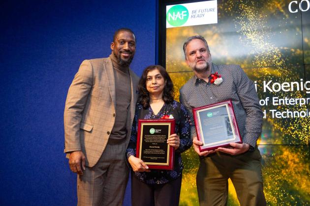 A man on the left poses with two award winners to his right on the stage.