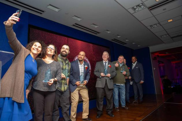 A woman on the left holds up her phone to take a selfie with a group of honorees on a stage.