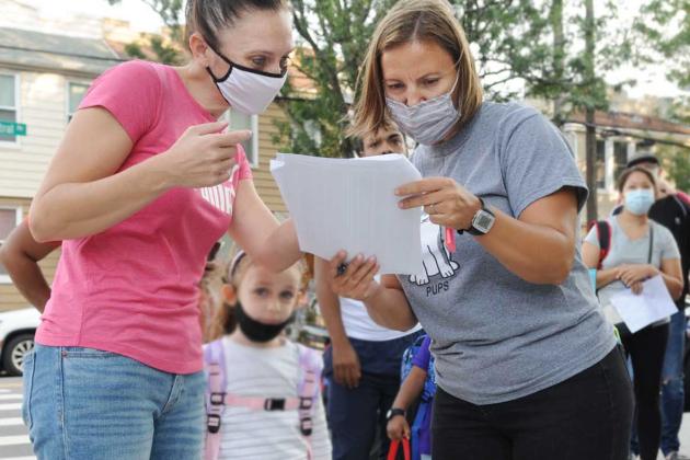 Two teachers both wearing masks look down at a piece of paper with a list of students. The teacher on the left is wearing a pink Puma shirt and a pink and black mask while the teacher next to her is wearing a grey t shirt and mask. 