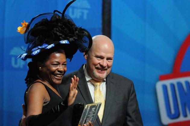 A woman wearing a large hat poses with a photo with a man on a stage
