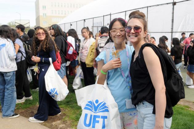 Two women with glasses pose for a photo amongst a crowd of many others in a field with a fence around it, one holding a UFT swag bag