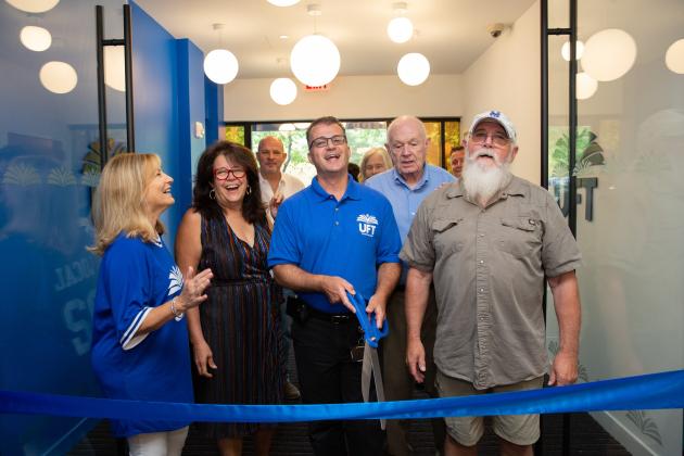 A group of people stand excitedly by a ribbon as a man prepares to cut it with large scissors