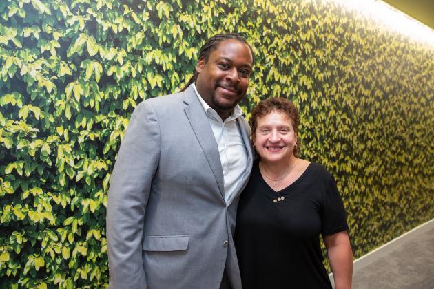 A man and woman pose for a photo against a wall painted with leaves.