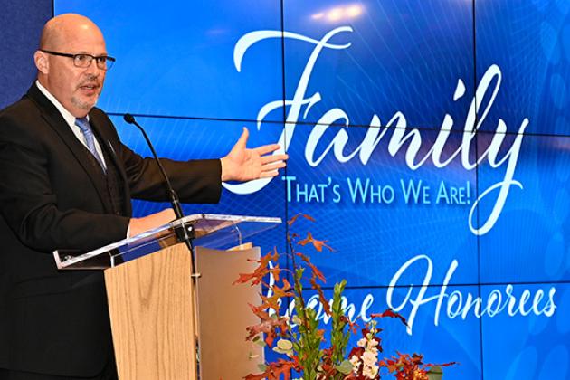 A man is standing at a podium speaking to an audience. A sign behind him reads "Family, that's what we are!"