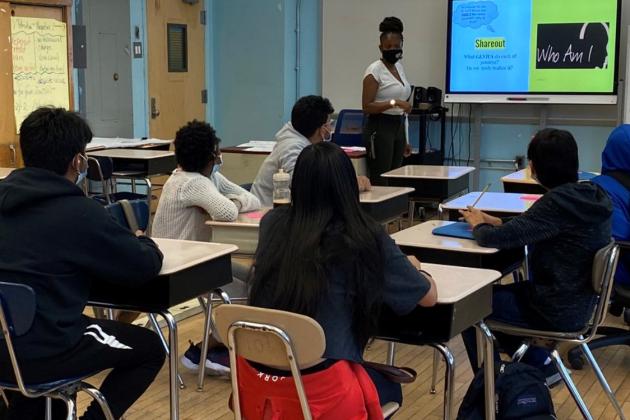 A teacher with a class of socially distanced students are engaged in a lesson. All are masked and the teacher is referencing material on the white board. 