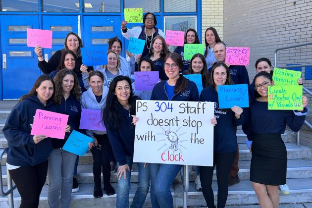 A group of teachers hold their signs in front of their school building.