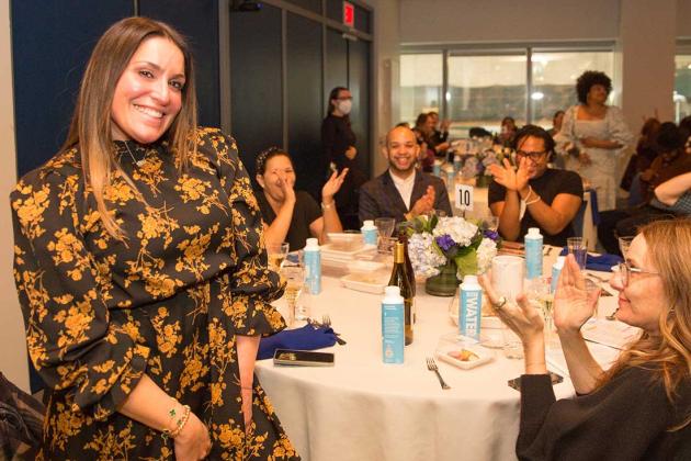 A woman poses and smiles while others at her table clap for her
