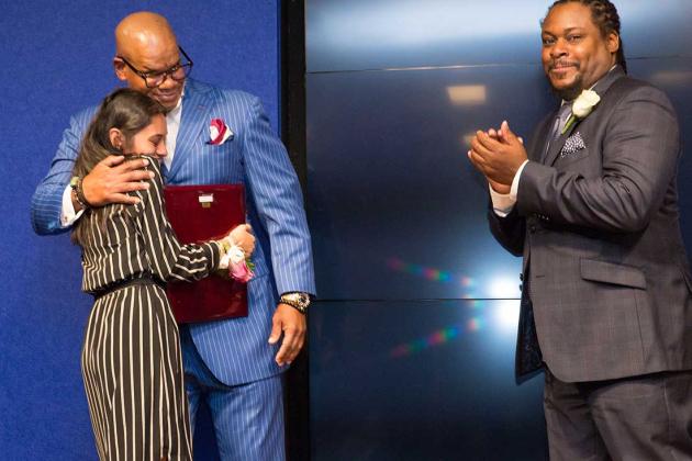 A woman hugs a bald man in a blue pinstripe suit while another man looks at the camera and claps 