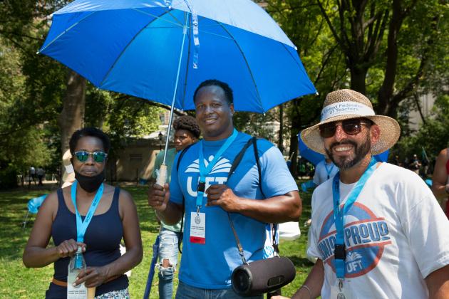 A woman on the left and two men, one with an umbrella smile for a picture in a park.