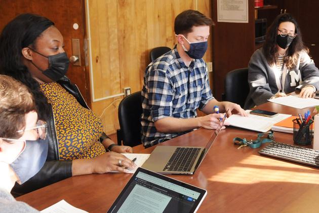 Masked people sitting at a large wooden conference table with laptops, pens and paper