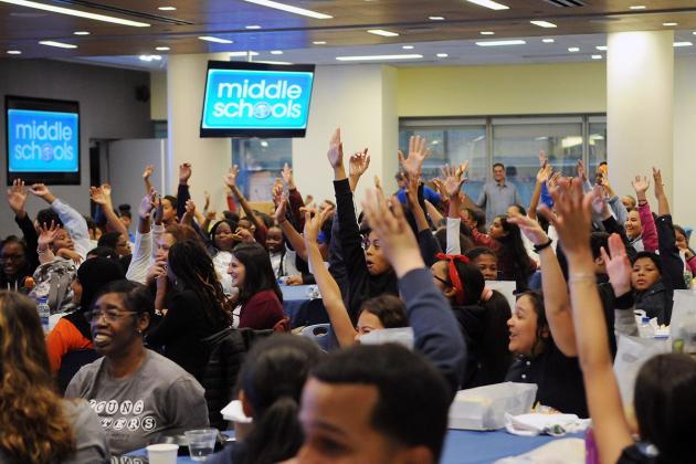 Large group of students sitting at tables with their hands in the air