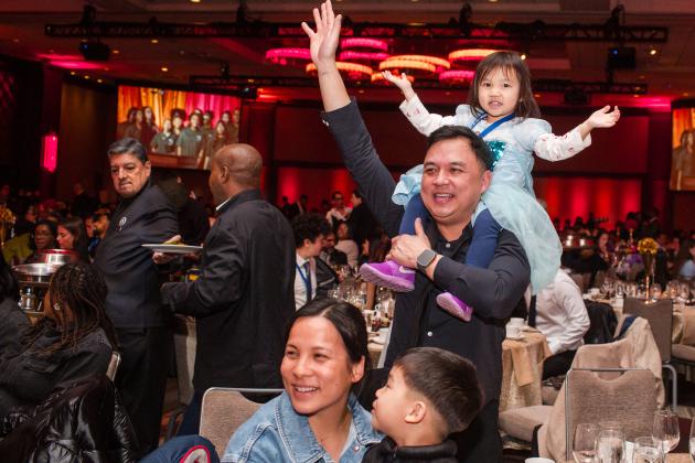 A man waves to the crowd with a child sitting on his shoulders. The man is being honored at the 2025 UFT Career and Technical Education Awards at the Sheraton New York Times Square Hotel on Feb. 26, 2025.