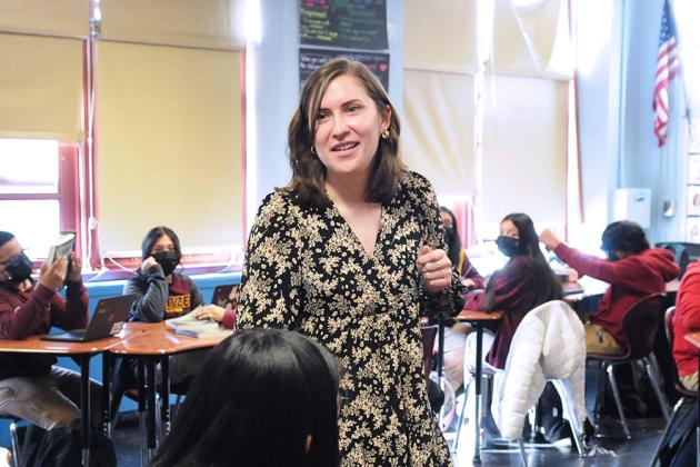Female teacher standing in room with others who are sitting.