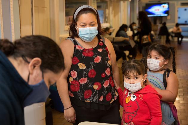 A woman and two young children wearing masks stand behind a table, looking down at something