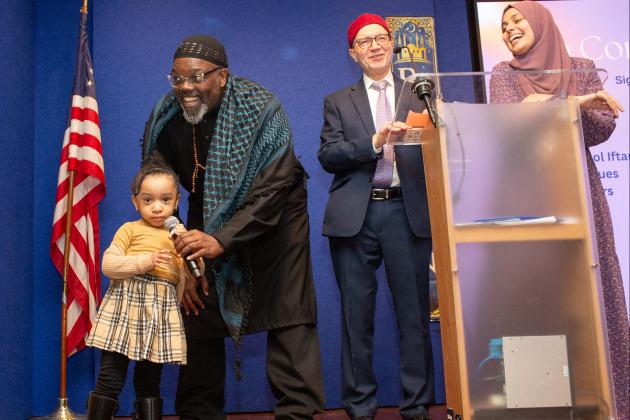 A child speaks into a microphone on stage as the adults look on and smile at the UFT Muslim Heritage Committee Iftar Dinner on March 7, 2025. 