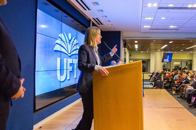 Blonde woman at podium on stage speaks to a seated crowd