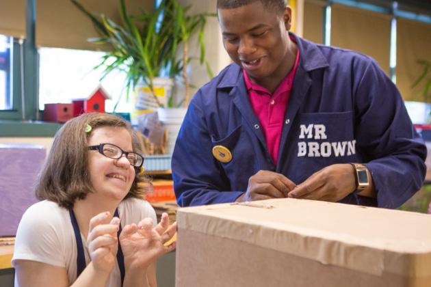 an educator in a jumpsuit that says 'Mr. Brown' assists a student who is laughing