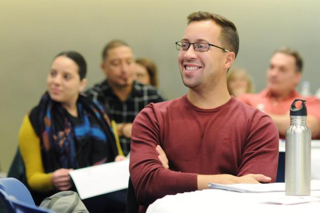 Smiling man with glasses in foreground sitting in classroom, other adults sitting in background