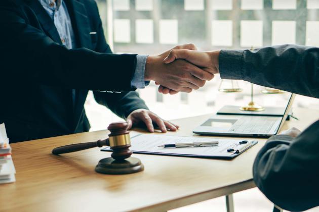Two men shaking hands across a desk after signing a document. There are papers and a gavel on the desk