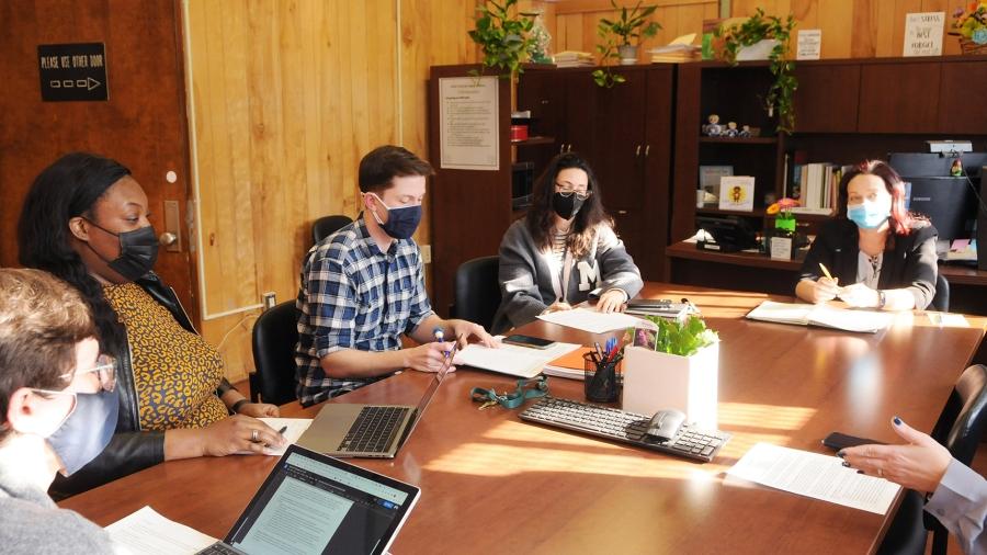 Masked people sitting at a large wooden conference table with laptops, pens and paper