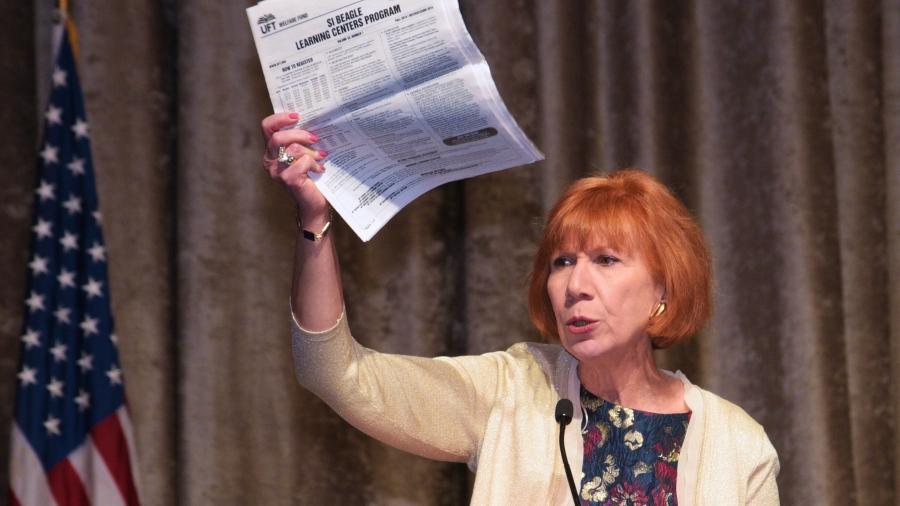 Mature red headed woman stands at podium holding a newspaper in hand