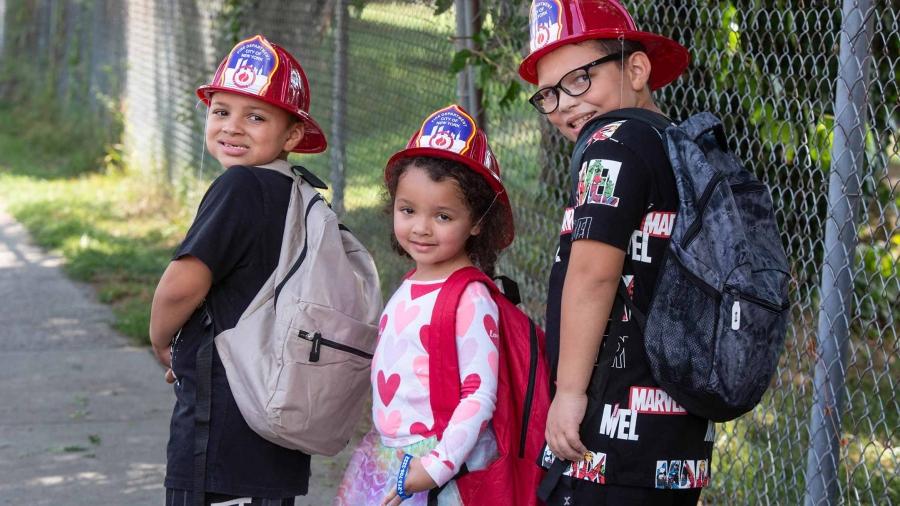 Three children wearing backpacks and fire helmets