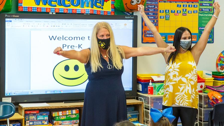 Two masked women stand with arms outstretched at the head of a classroom in front of an electronic board reading "welcome to pre-k"