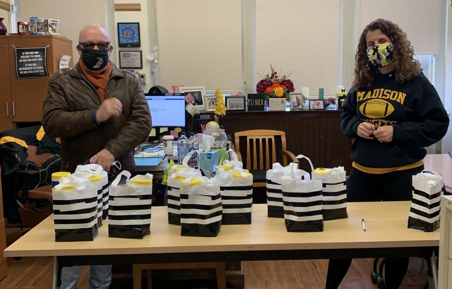A man and a woman, both wearing masks, stand behind a table in a public school office full of bags to distribute to share with members of the school community.