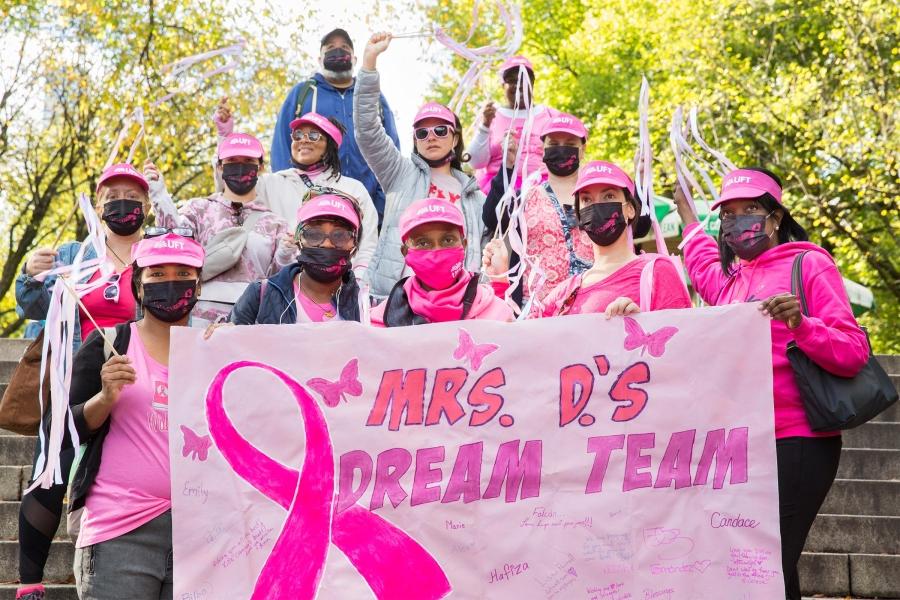 Twelve UFT members wearing pink regalia to honor people with Breast cancer stand together, holding streamers and a sign reading "Mrs. D's Dream Team"