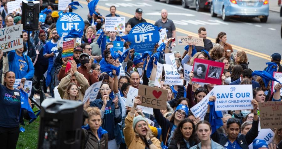 A large crowd gathers on the street, holding signs supporting the UFT and the DOE's Division of Early Childhood Education
