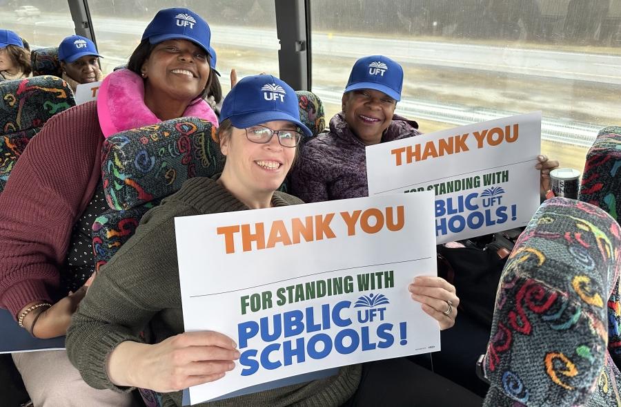 Three women wearing UFT hats pose together on a bus for a photo. Two are holding signs reading "Thank you for standing with Public Schools." 