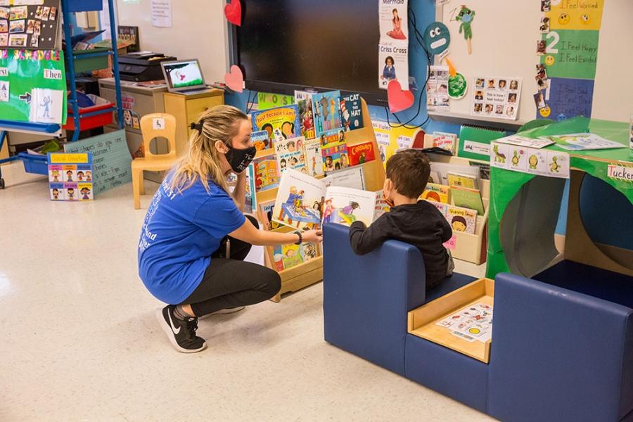 A teacher in a blue t-shirt crouches down to the eye level of one of her students so they can read the picture book she is holding. 