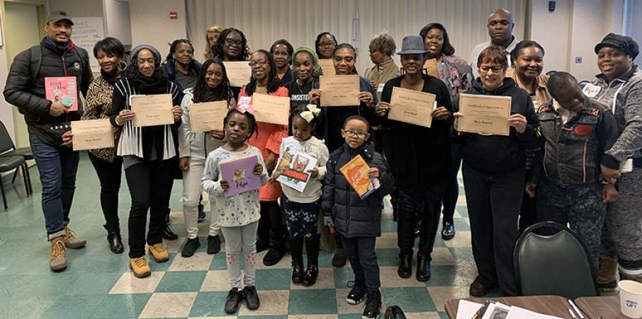 A group of about 25 adults and children hold up certificates, smiling.