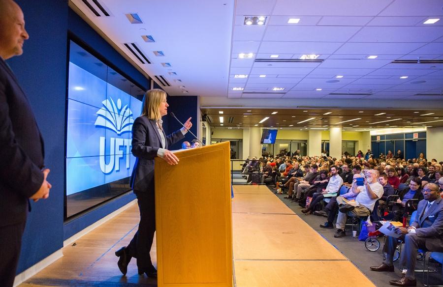 Blonde woman at podium on stage speaks to a seated crowd